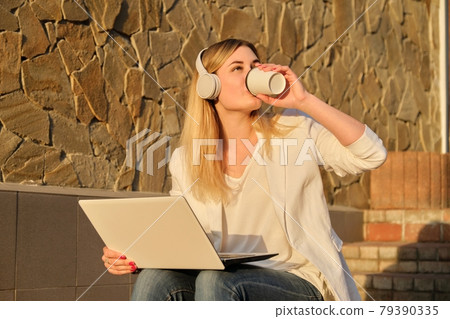 Young beautiful female in headphones with laptop drinking coffee in paper cup Young beautiful female in headphones with laptop drinking coffee in paper cup 79390335