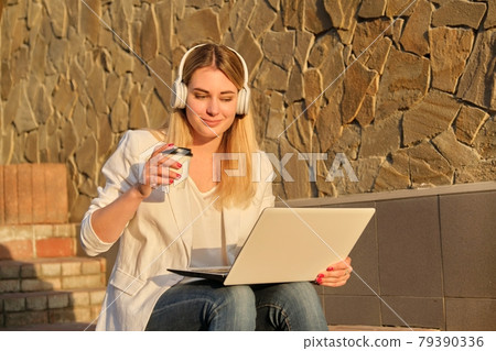 Young beautiful female in headphones with laptop drinking coffee in paper cup Young beautiful female in headphones with laptop drinking coffee in paper cup 79390336