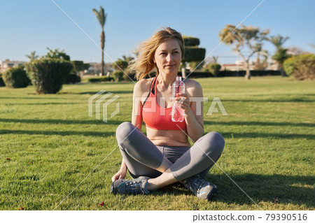 Mature woman doing morning exercises on green grass in park, with bottle of water Mature woman doing morning exercises on green grass in park, with bottle of water 79390516