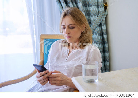 Serious middle aged woman with smartphone sitting on chair at home Serious middle aged woman with smartphone sitting on chair at home 79391904