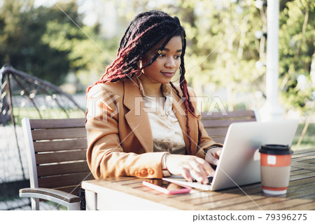 Young smiling african american woman sitting at the table in street cafe and using laptop Young smiling african american woman sitting at the table in street cafe and using laptop 79396275