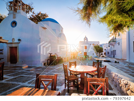 Sunrise in alleyways of Mykonos, Greece. Greek whitewashed churches in small town square, street cafe tables and chairs, sun and lens flares. Sunrise in alleyways of Mykonos, Greece. Greek whitewashed churches in small town square, street cafe tables and chairs, sun and lens flares. 79397642