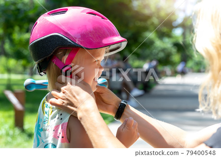 Close-up mom parent hand put on and fasten safety helmet on cute blond caucasian toddler girl for riding bike or scooter city street park outdoors on summer day. Child sport activity protection care 79400548