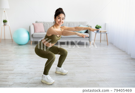 Full length portrait of happy young Indian woman doing squats exercises at home, blank space Full length portrait of happy young Indian woman doing squats exercises at home, blank space 79405469