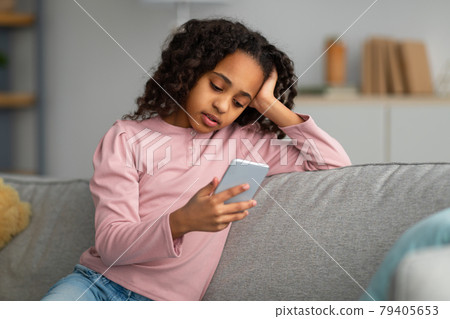 Bored African American Teen Girl Using Smartphone Browsing Internet Sitting On Sofa At Home. Modern Boring Childhood 79405653