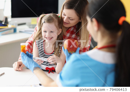 Doctor pediatrician holding bottle with medical capsules and red apple in front of little girl and mom in clinic 79406413
