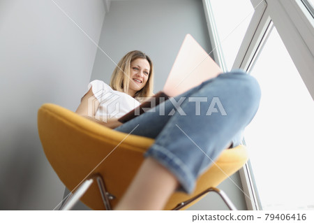 Young smiling woman sitting in yellow armchair and looking at laptop screen Young smiling woman sitting in yellow armchair and looking at laptop screen 79406416