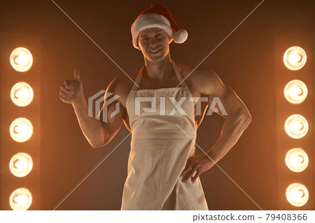 Hot Christmas dessert. Muscular man wearing apron and Santa hat showing thumb up gesture and smiling at camera, lamps illumination on background Hot Christmas dessert. Muscular man wearing apron and Santa hat showing thumb up gesture and smiling at camera, lamps illumination on background 79408366