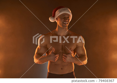 Portrait of muscular man wearing Christmas Santa hat, showing thumb up on smoky background 79408367