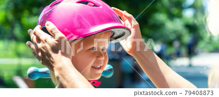Close-up mom parent hand put on and fasten safety helmet on cute blond caucasian toddler girl for riding bike or scooter city street park outdoors on summer day. Child sport activity protection care 79408873