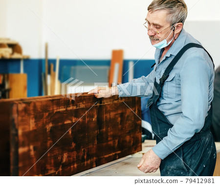 A gray-haired carpenter with glasses works with wood on a workbench in a carpentry shop. A gray-haired carpenter with glasses works with wood on a workbench in a carpentry shop. 79412981