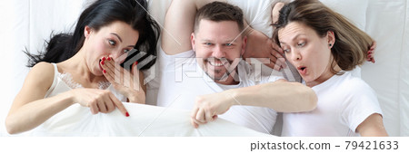 Man lying with two lovers in bed. Women looking under cover and wondering 79421633