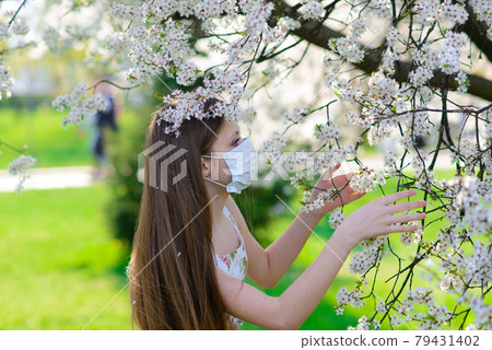 Lonely teenage girl wearing protection medicine mask against virus and allergy enjoying spring flower blossom in blooming garden Lonely teenage girl wearing protection medicine mask against virus and allergy enjoying spring flower blossom in blooming garden 79431402