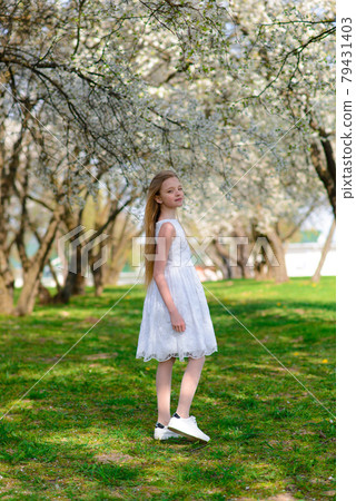 Beautiful blue-eyed girl with blond hair in white dress walking in flower garden,emotional photo. 79431403