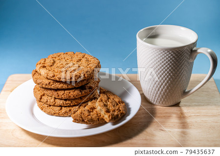 A mug with milk and oatmeal cookies on a wooden table. White plate with fresh oatmeal cookies on a blue background. A mug with milk and oatmeal cookies on a wooden table. White plate with fresh oatmeal cookies on a blue background. 79435637