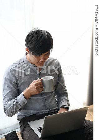 Portrait of young businessman working on laptop while drinking coffee and sitting on wooden floor. Portrait of young businessman working on laptop while drinking coffee and sitting on wooden floor. 79441913