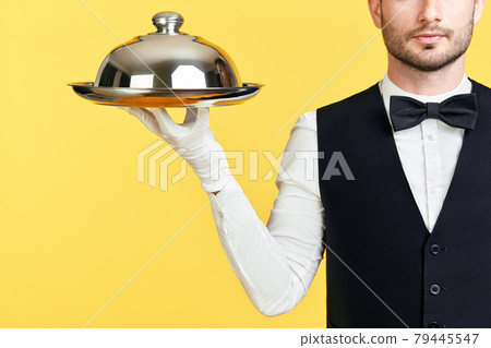 Handsome elegant waiter holding tray and cloche ready to serve on yellow background 79445547
