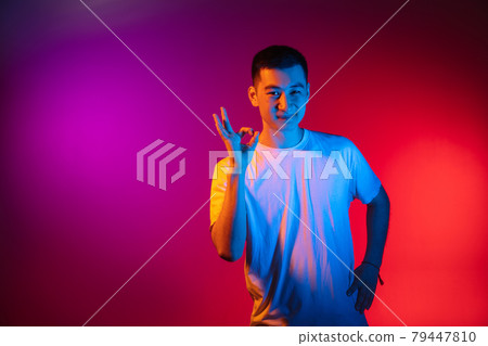 One Asian young man isolated on studio background in gradient pink purple neon light, colour filter. Concept of human emotions, facial expression. 79447810