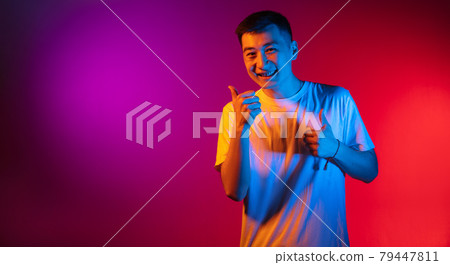 One Asian young man isolated on studio background in gradient pink purple neon light, colour filter. Concept of human emotions, facial expression. One Asian young man isolated on studio background in gradient pink purple neon light, colour filter. Concept of human emotions, facial expression. 79447811