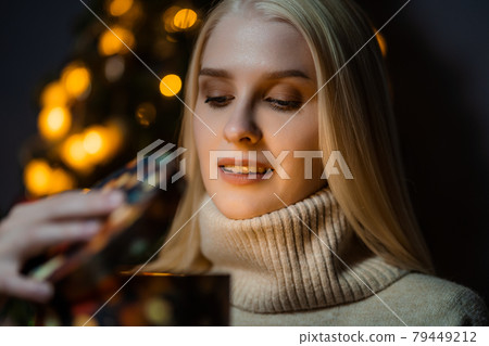 A cute woman in a cozy sweater opens a New Year's gift against the background of a Christmas tree. Garland lights are burning in the background. 79449212