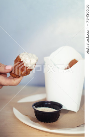 Close-up of chicken wings fried in batter. In a fast food restaurant, there is a cardboard box of chicken and sauce on the table. Sun rays fall beautifully on the hand of the girl who holds the food Close-up of chicken wings fried in batter. In a fast food restaurant, there is a cardboard box of chicken and sauce on the table. Sun rays fall beautifully on the hand of the girl who holds the food 79450516