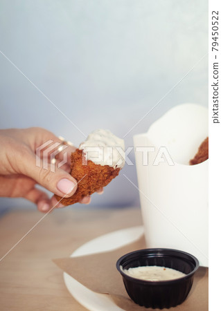 Close-up of chicken wings fried in batter. In a fast food restaurant, there is a cardboard box of chicken and sauce on the table. Sun rays fall beautifully on the hand of the girl who holds the food Close-up of chicken wings fried in batter. In a fast food restaurant, there is a cardboard box of chicken and sauce on the table. Sun rays fall beautifully on the hand of the girl who holds the food 79450522