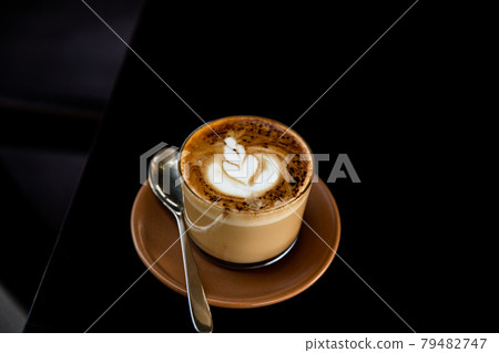 Glass of cappuccino with latte art on saucer and with spoon on the black table Glass of cappuccino with latte art on saucer and with spoon on the black table 79482747