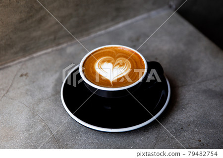 Black cup of cappuccino with latte art of heart shape on saucer on concrete background. Black cup of cappuccino with latte art of heart shape on saucer on concrete background. 79482754