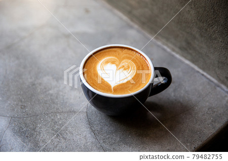 Black cup of cappuccino with latte art of heart shape on saucer on concrete background. Black cup of cappuccino with latte art of heart shape on saucer on concrete background. 79482755