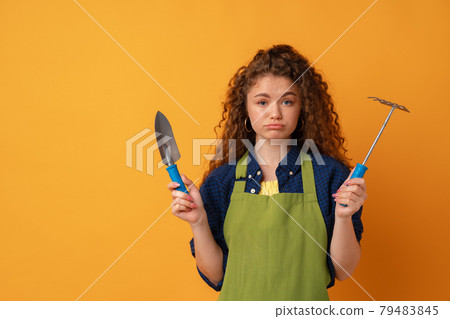 Young gardener woman wearing apron holding gardening tools against yellow background Young gardener woman wearing apron holding gardening tools against yellow background 79483845