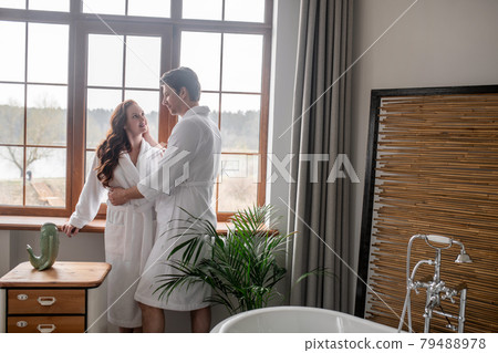 Two people in white bathrobes standing near the window and looking affectionate 79488978
