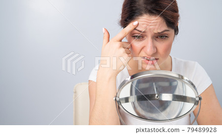 Caucasian woman looks at herself in the mirror and is upset because of facial wrinkles on her face. Caucasian woman looks at herself in the mirror and is upset because of facial wrinkles on her face. 79489928