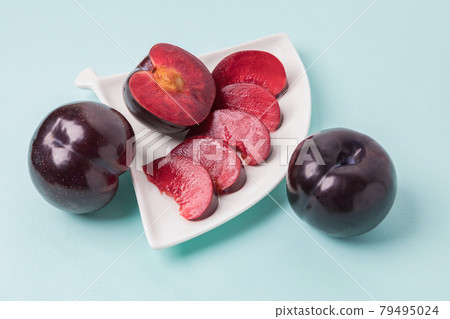 On a white saucer there is a large plum cut into wedges, next to it there are two more plums. On a white saucer there is a large plum cut into wedges, next to it there are two more plums. 79495024
