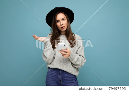 Beautiful young asking dissatisfied brunette woman wearing black hat and grey sweater holding 79497293