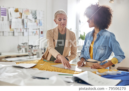 Mature lady boss checks sewing patterns made by young African-American seamstress at cutting table Mature lady boss checks sewing patterns made by young African-American seamstress at cutting table 79506270