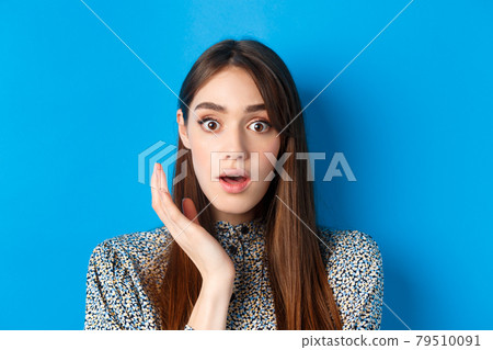 Candid girl with long hair and makeup, looking shocked and surprised, open mouth and stare speechless at camera, standing on blue background 79510091