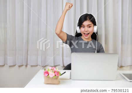Young smart and active asian female student sitting at table with headset, using computer and taking note for studying online with exciting emotion (new normal concept) Young smart and active asian female student sitting at table with headset, using computer and taking note for studying online with exciting emotion (new normal concept) 79530121