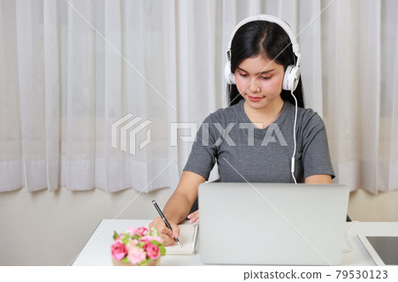 Young smart and active asian female student sitting at table with headset, using computer and taking note for studying online with happy smiling face (new normal concept) Young smart and active asian female student sitting at table with headset, using computer and taking note for studying online with happy smiling face (new normal concept) 79530123