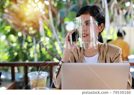 Young asian woman in casual dress with face shield for healthcare, sitting in coffee shop and using on smartphone and working on laptop computer. New normal and social distancing concept 79530180