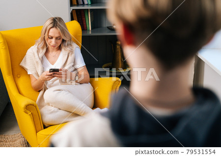 Close-up rear view of teen Caucasian kid son looking at mother sitting on yellow armchair in cozy living room. 79531554