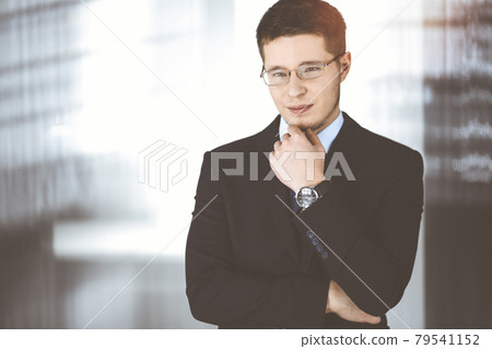 Young businessman wearing suit, with glasses, while standing in the sunny office. Business success concept nowadays 79541152
