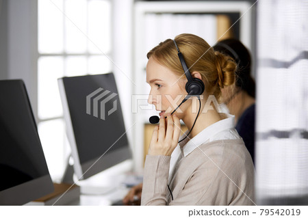 Friendly businesswoman talking by headset while sitting in office. Call center and diverse people group in business 79542019