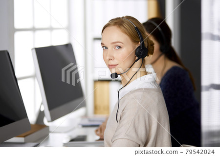 Friendly businesswoman talking by headset while sitting in office. Call center and diverse people group in business 79542024