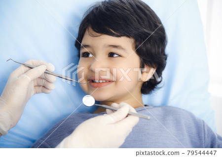 Cute arab boy sitting at dental chair with open mouth during oral checking up with doctor. Visiting dentist office. Stomatology concept Cute arab boy sitting at dental chair with open mouth during oral checking up with doctor. Visiting dentist office. Stomatology concept 79544447