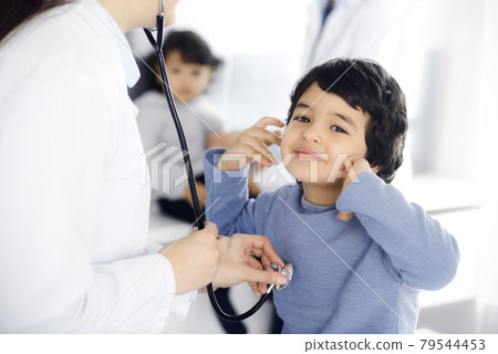 Doctor-woman examining a child patient by stethoscope. Cute arab boy and his brother at physician appointment. Medicine concept Doctor-woman examining a child patient by stethoscope. Cute arab boy and his brother at physician appointment. Medicine concept 79544453