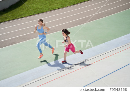 Friends in sports suits running on a treadmill at stadium and looking at the each other 79548568