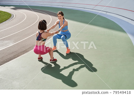 Women looking at the each other while lunge and squat on the grass in the stadium 79549418