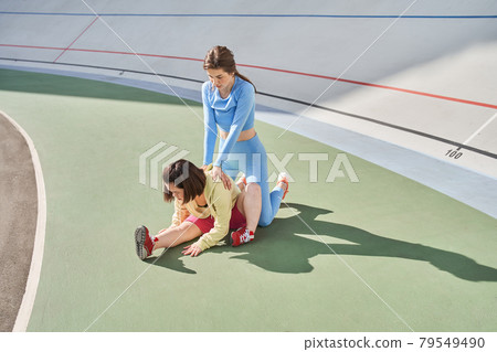 Woman helping to stretching to her midget friend or client while being at the stadium Woman helping to stretching to her midget friend or client while being at the stadium 79549490