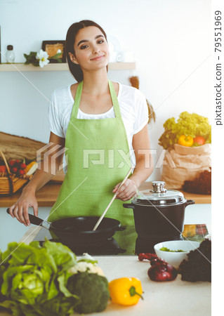 An attractive young dark-haired woman frying meal at worm pan by new keto recipe while standing and smiling in sunny kitchen. Cooking and householding concepts 79551969