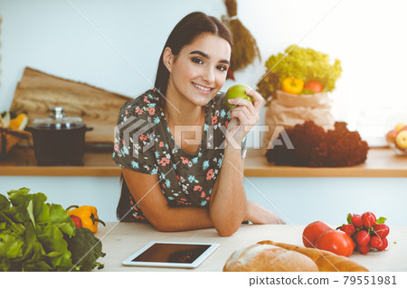 An attractive young dark-haired woman holding green apple and choosing the recipe for a delicious meal while smiling in sunny kitchen. Tablet pc is the best cookbook 79551981